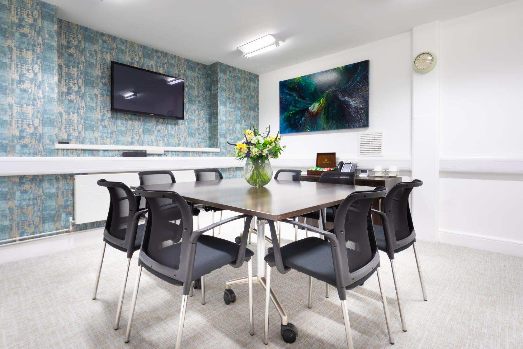 A meeting room with a rectangular table, black chairs, wall-mounted TV, and floral centerpiece at Landmark: Marylebone, London.