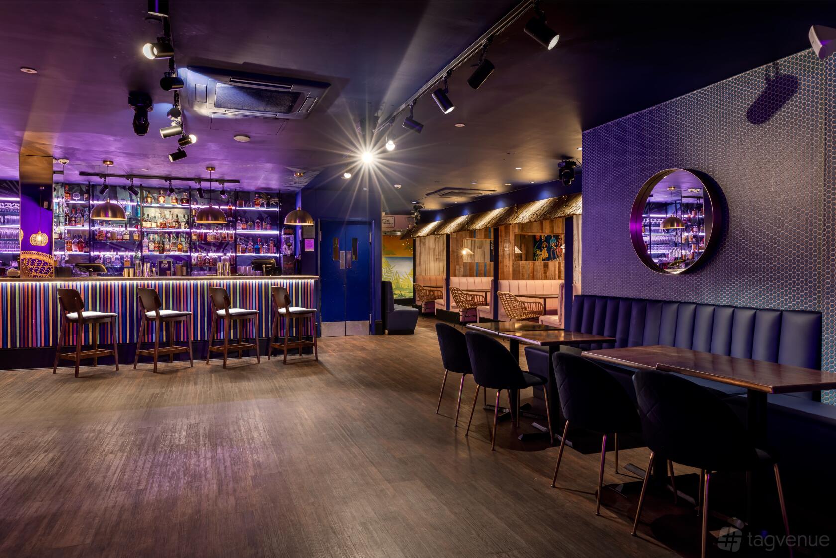 A function room with a striped-lit bar, velvet chairs, and wall mirrors at Cottons Shoreditch.