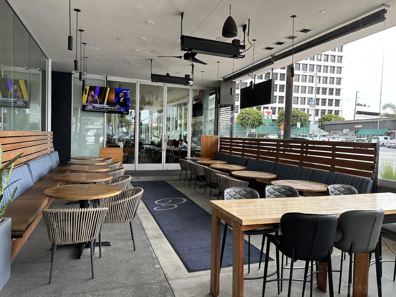 An outdoor restaurant patio with round wooden tables, wicker chairs, and bench seating at Pita Bar + Grill.