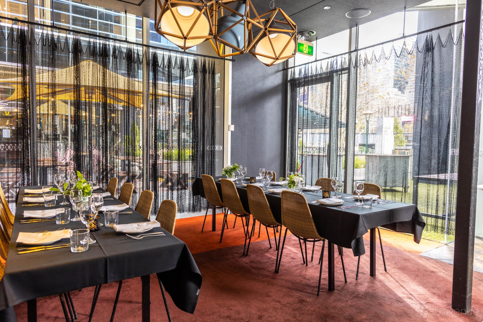 A dining room with floor-to-ceiling windows, geometric pendant lights, and tables set with black linens at Ludlow Bar.