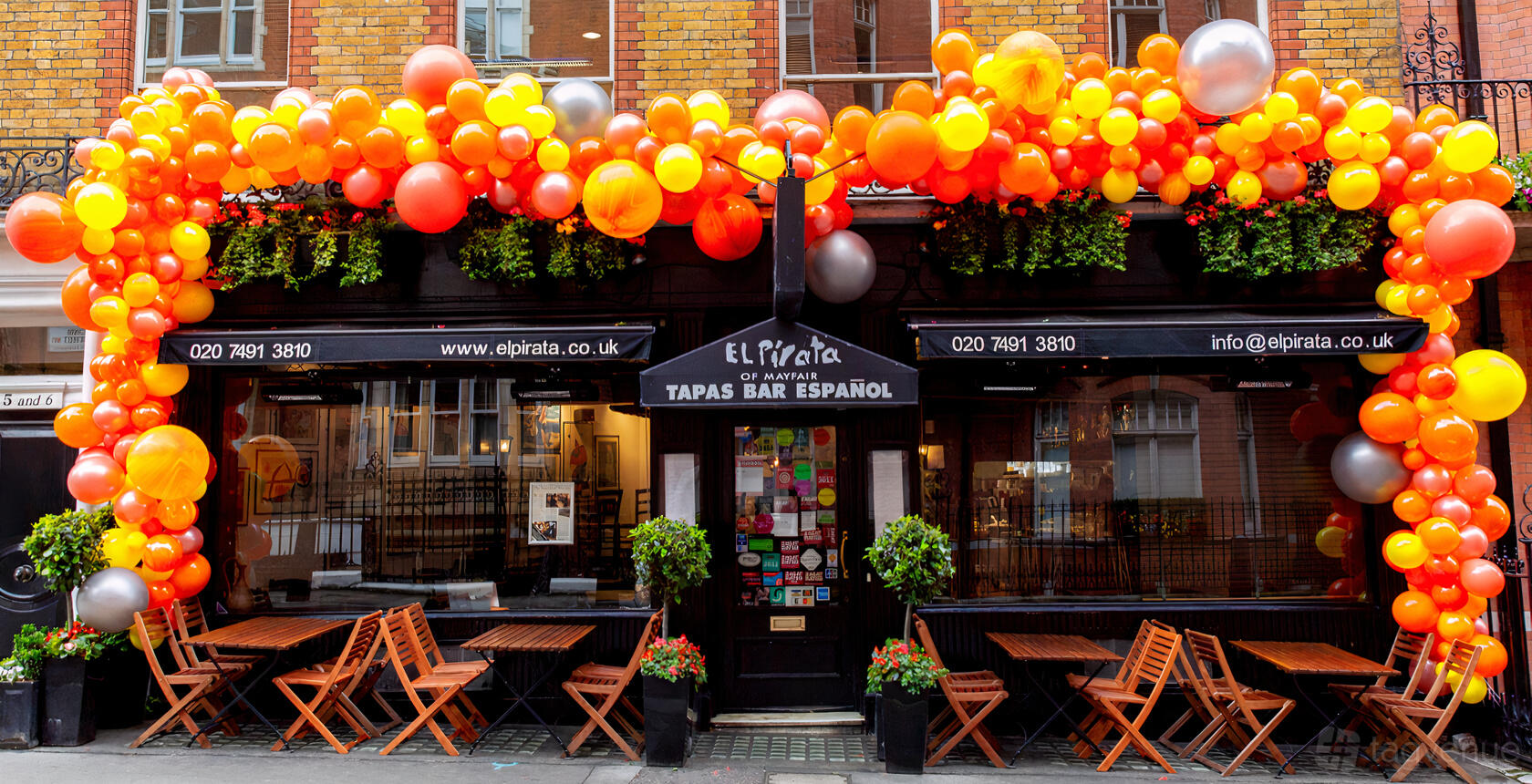 A restaurant with outdoor wooden tables, orange and red balloons, and brick exterior at El Pirata of Mayfair.