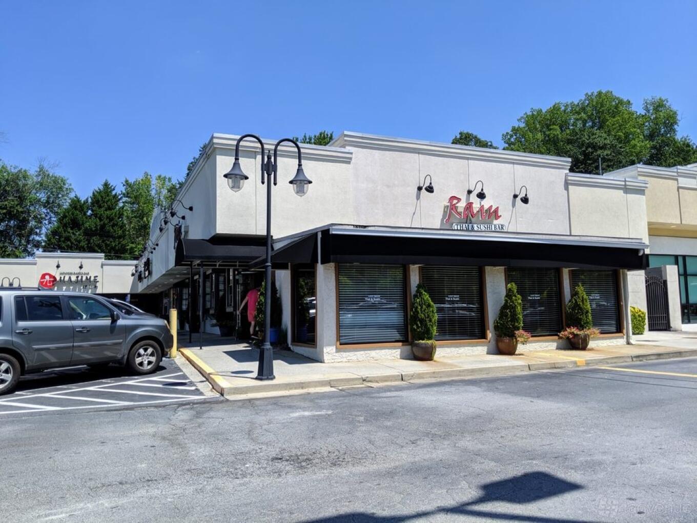 A restaurant with a light-colored facade, black awning, and large front windows at Rain Thai & Sushi Bar.