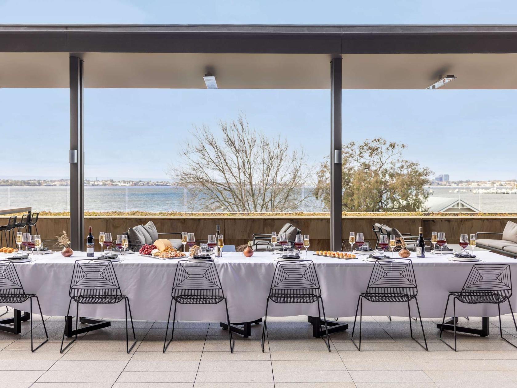 A restaurant terrace with a long table set with white linens, black chairs, and river views at Wonil Hotel Perth - Handwritten Collection.