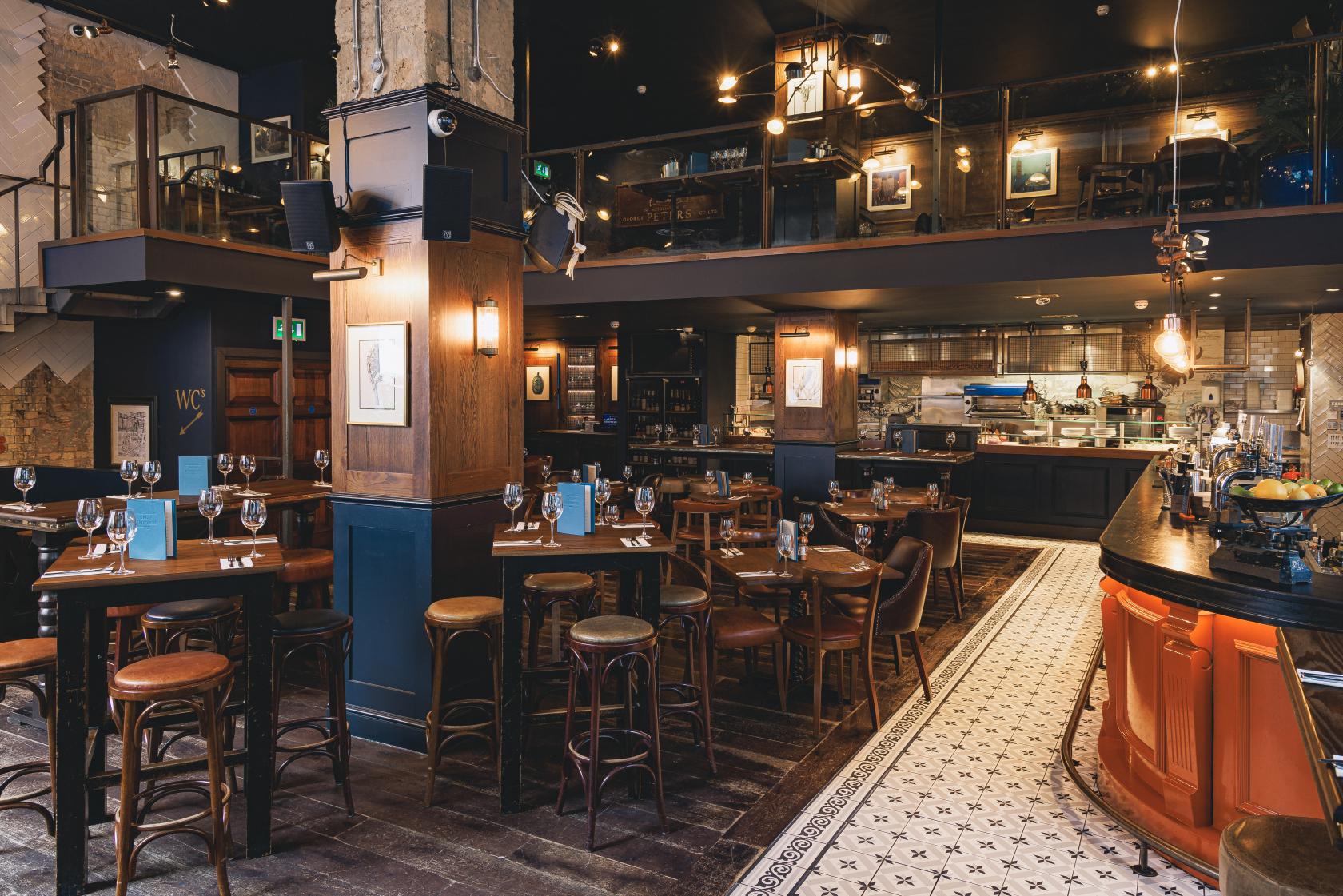 A bar with wooden tables, stools, exposed brick columns, and an open kitchen at Balls Brothers Austin Friars.