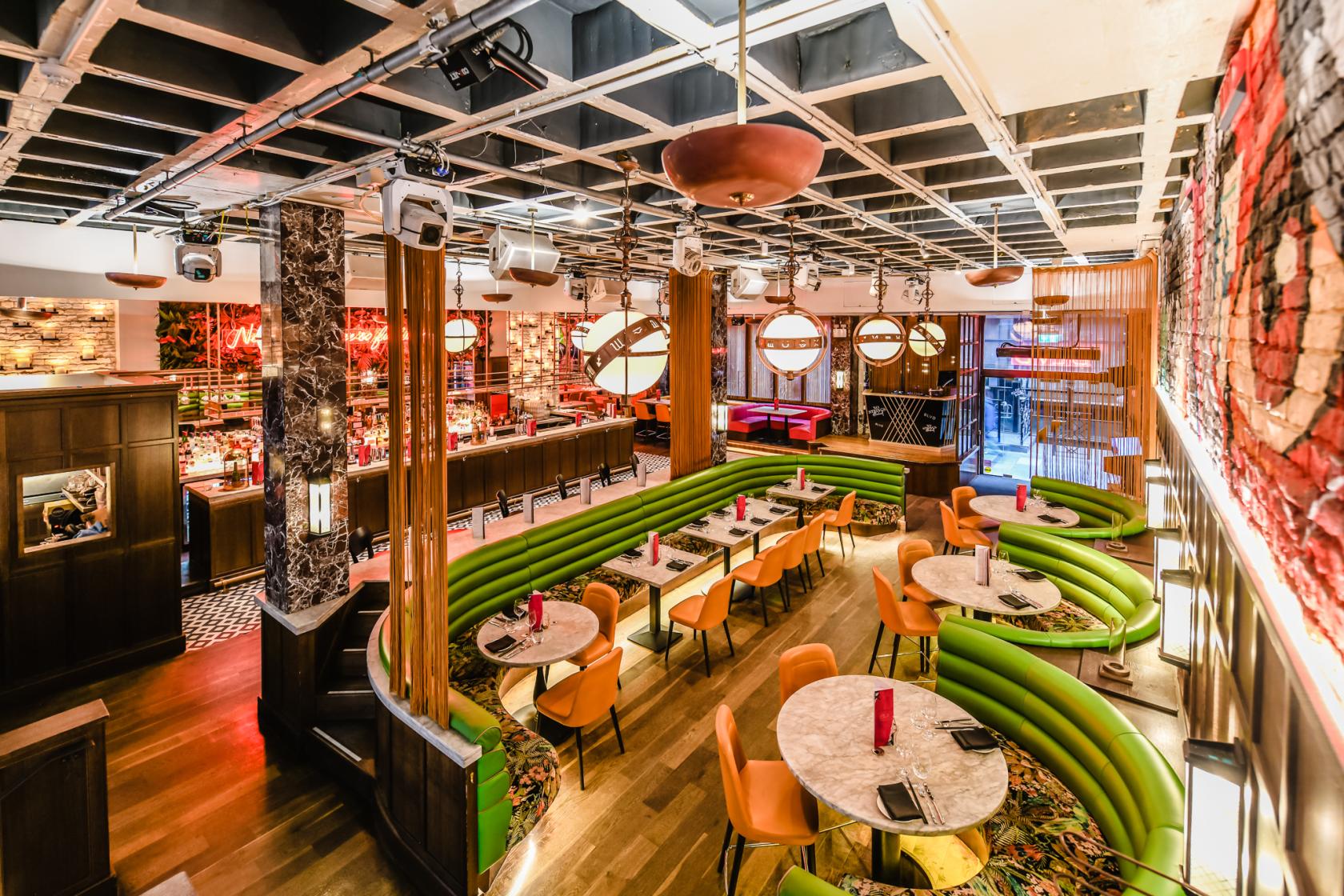 A bar with green velvet banquette seating, round marble tables, and a backlit counter at BLVD Manchester.