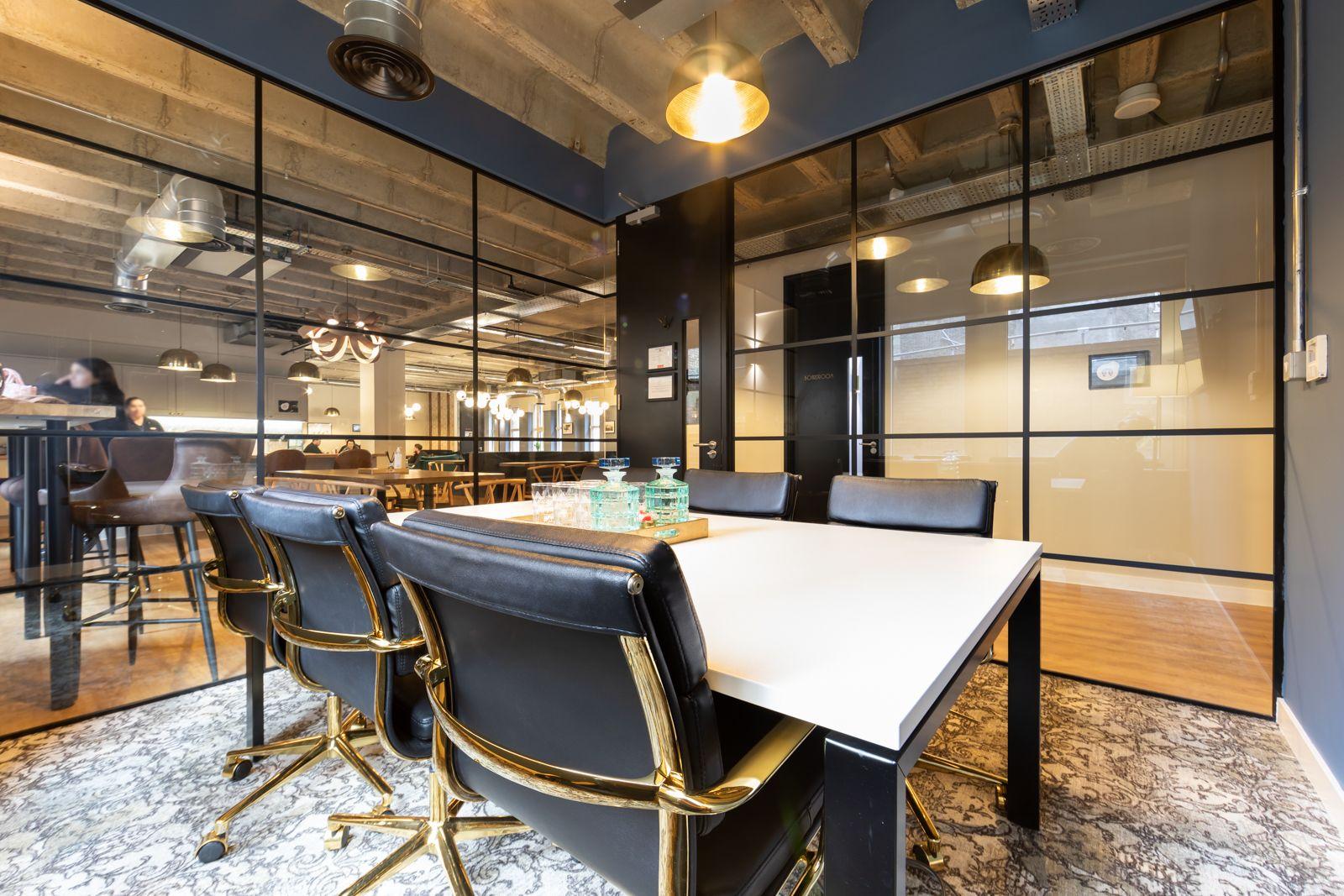 A meeting room with a white table, black chairs with gold trim, glass walls, and exposed ceiling at The Space Liverpool St.