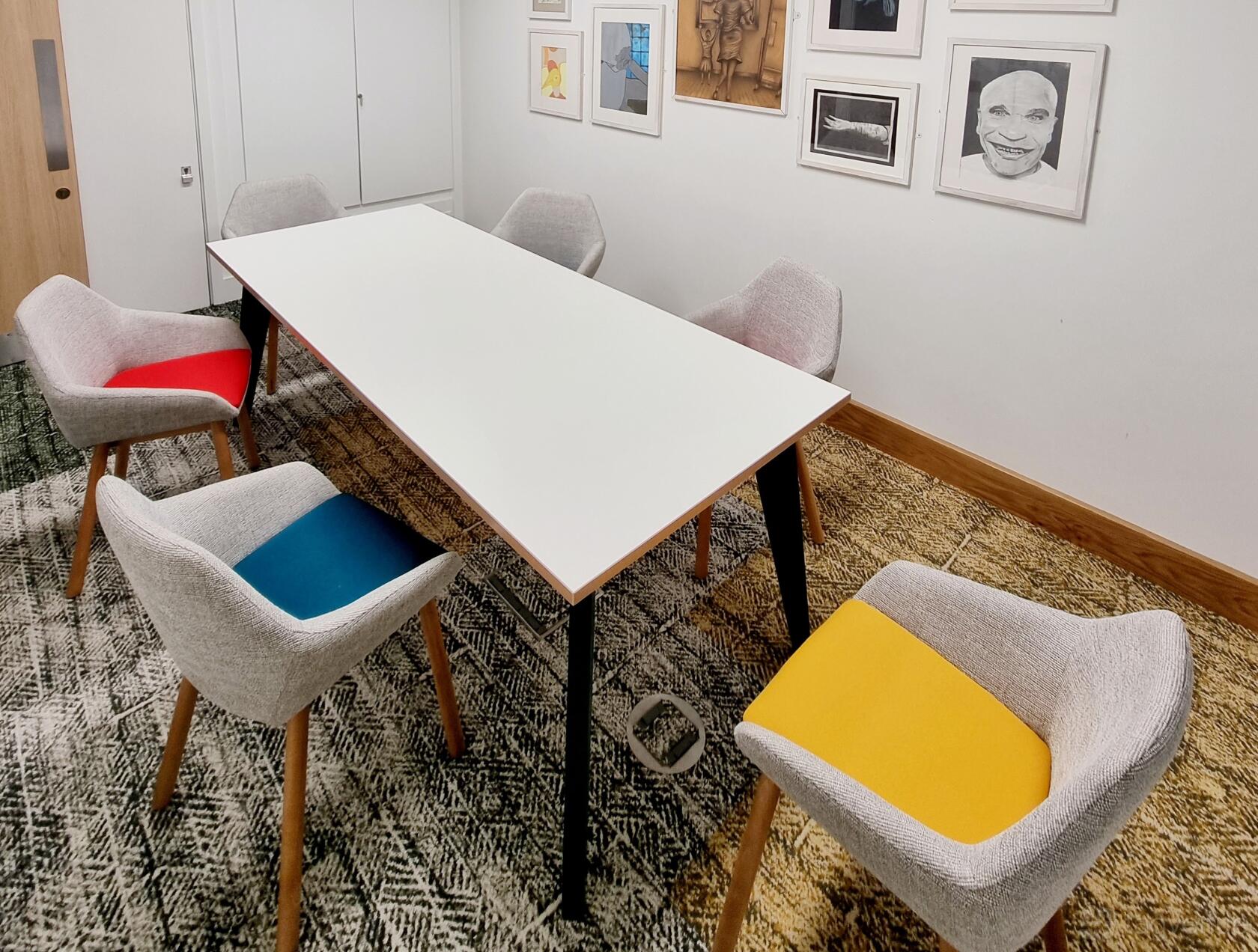 A meeting room with a rectangular white table, six upholstered chairs with colorful seats, and framed art at The Nest.