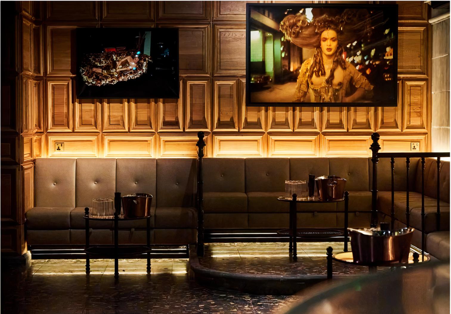 A nightclub lounge with tufted leather seating, wood-paneled walls, and large screens at BEAT LONDON.