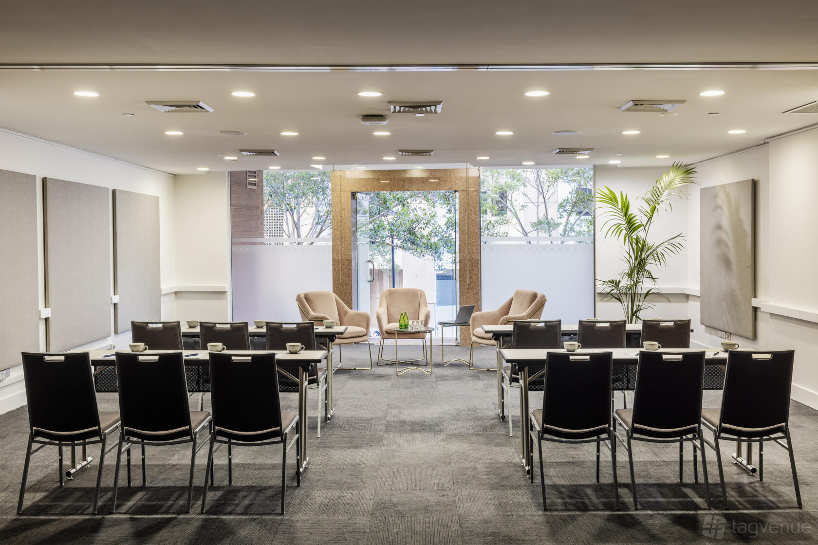 A function room with rows of black chairs, a small stage area with armchairs, and large windows at Sebel Quay West Suites Sydney.