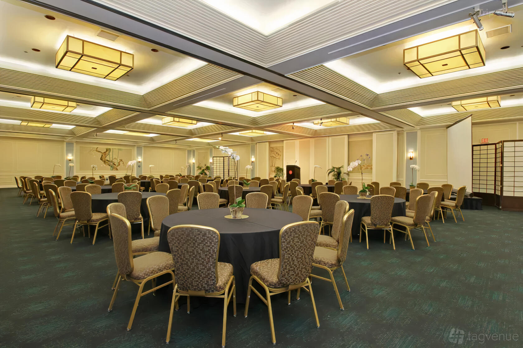 A hotel ballroom with round tables, upholstered chairs, decorative wall panels, and geometric ceiling lights at Hotel Kabuki.