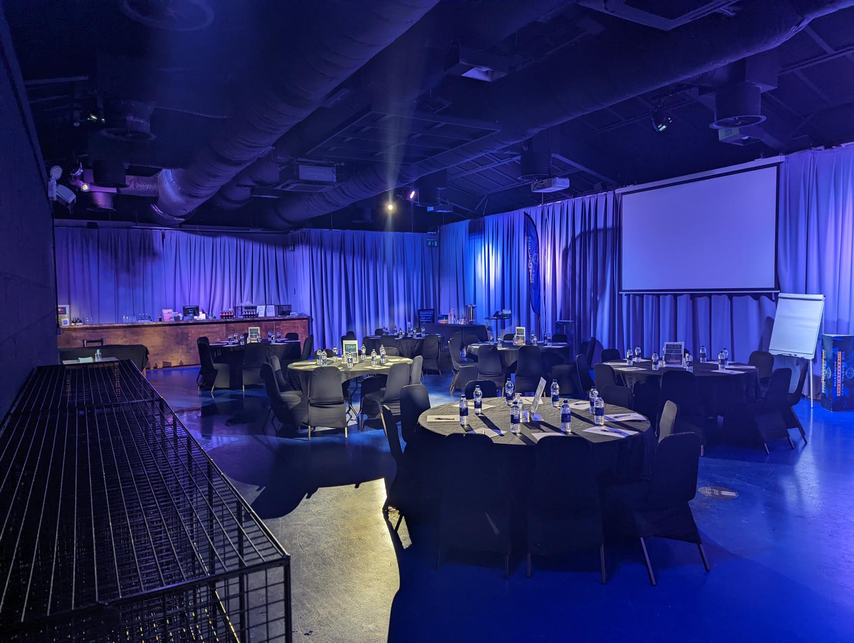 An entertainment centre with round tables, black chairs, and blue uplighting at The Crystal Maze LIVE Experience.