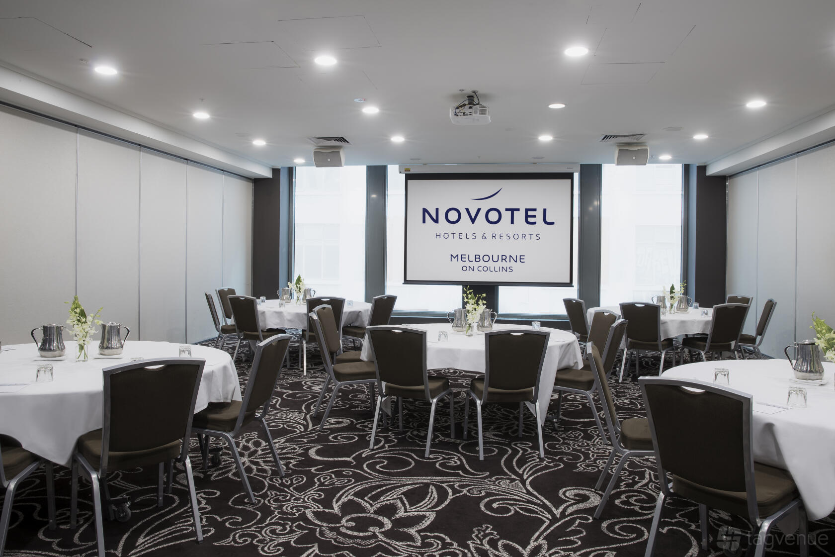 A function room with round tables covered in white linens, brown chairs, and a projector screen at Novotel Melbourne on Collins.