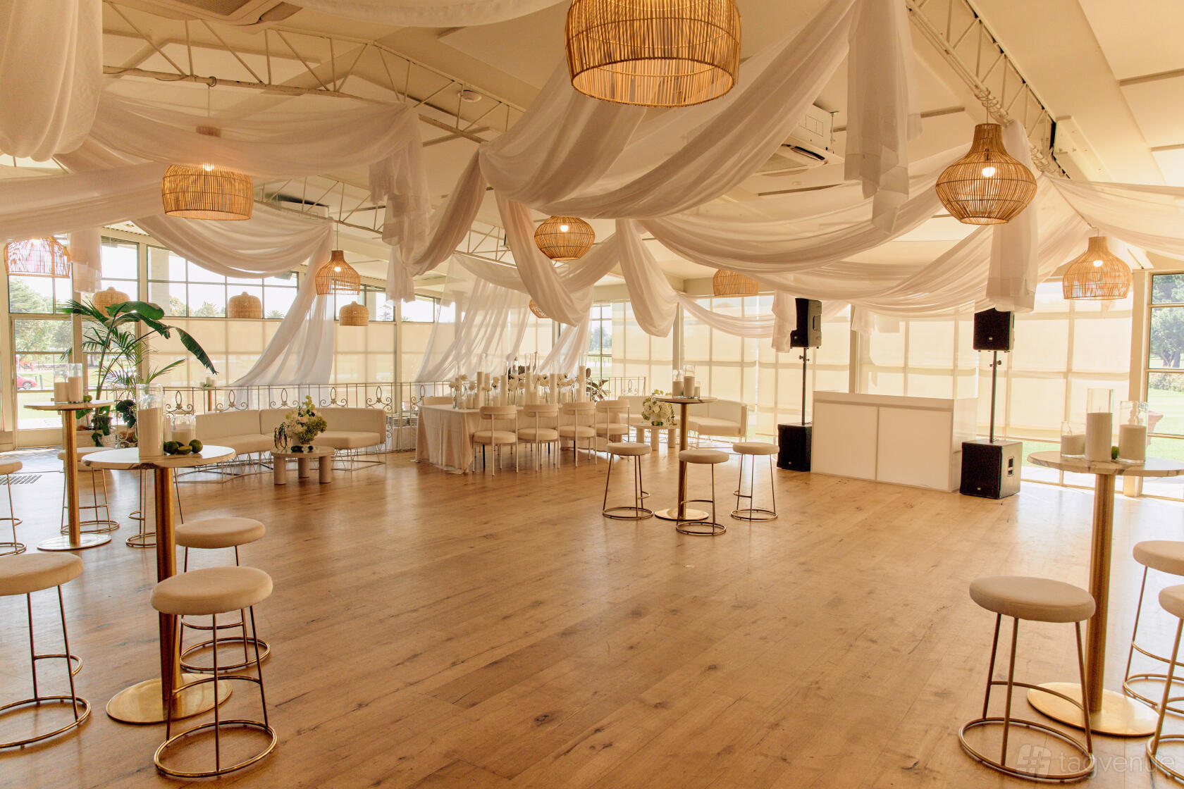 An event venue with wood flooring, draped white fabric ceiling, wicker pendant lights, and tall windows at Greenfields Albert Park.