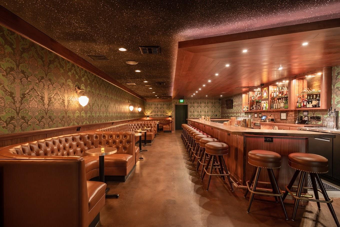 A bar with tufted leather banquettes, patterned wallpaper, and a wooden bar counter at The Grasshopper.