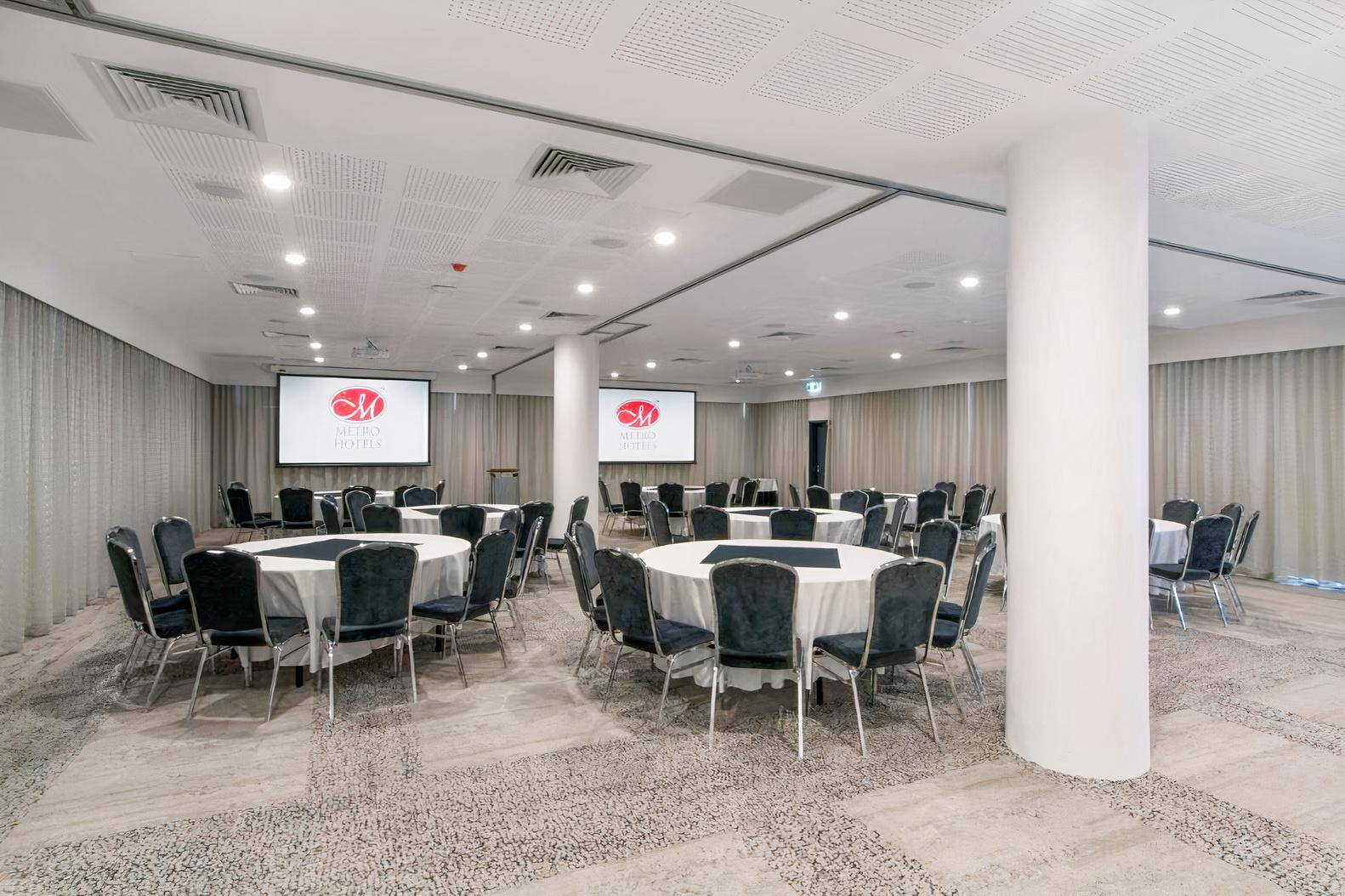 An event space with round tables, black chairs, and dual projection screens at Metro Hotel South Perth’s Swan Room.