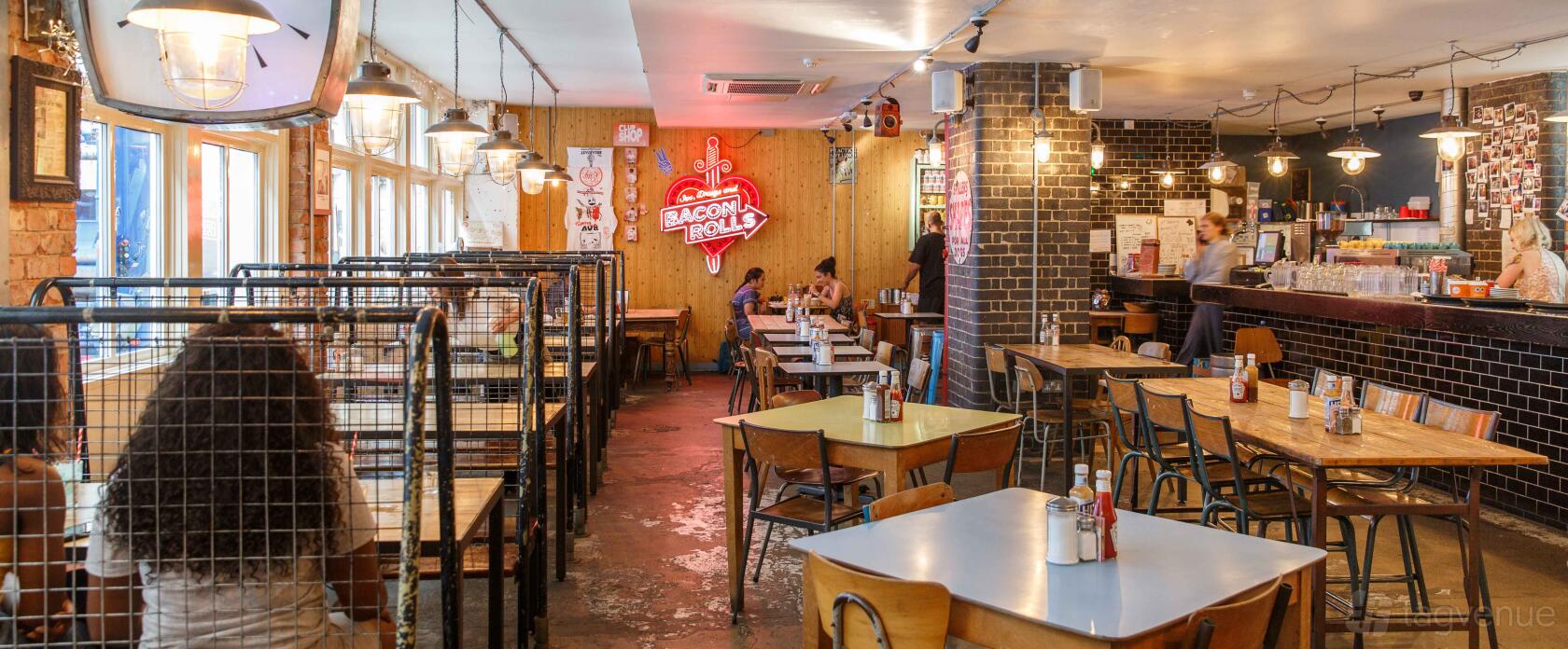 A cocktail bar with wooden tables, metal chairs, a neon bacon rolls sign, and exposed brick column at The Breakfast Club Spitalfields.