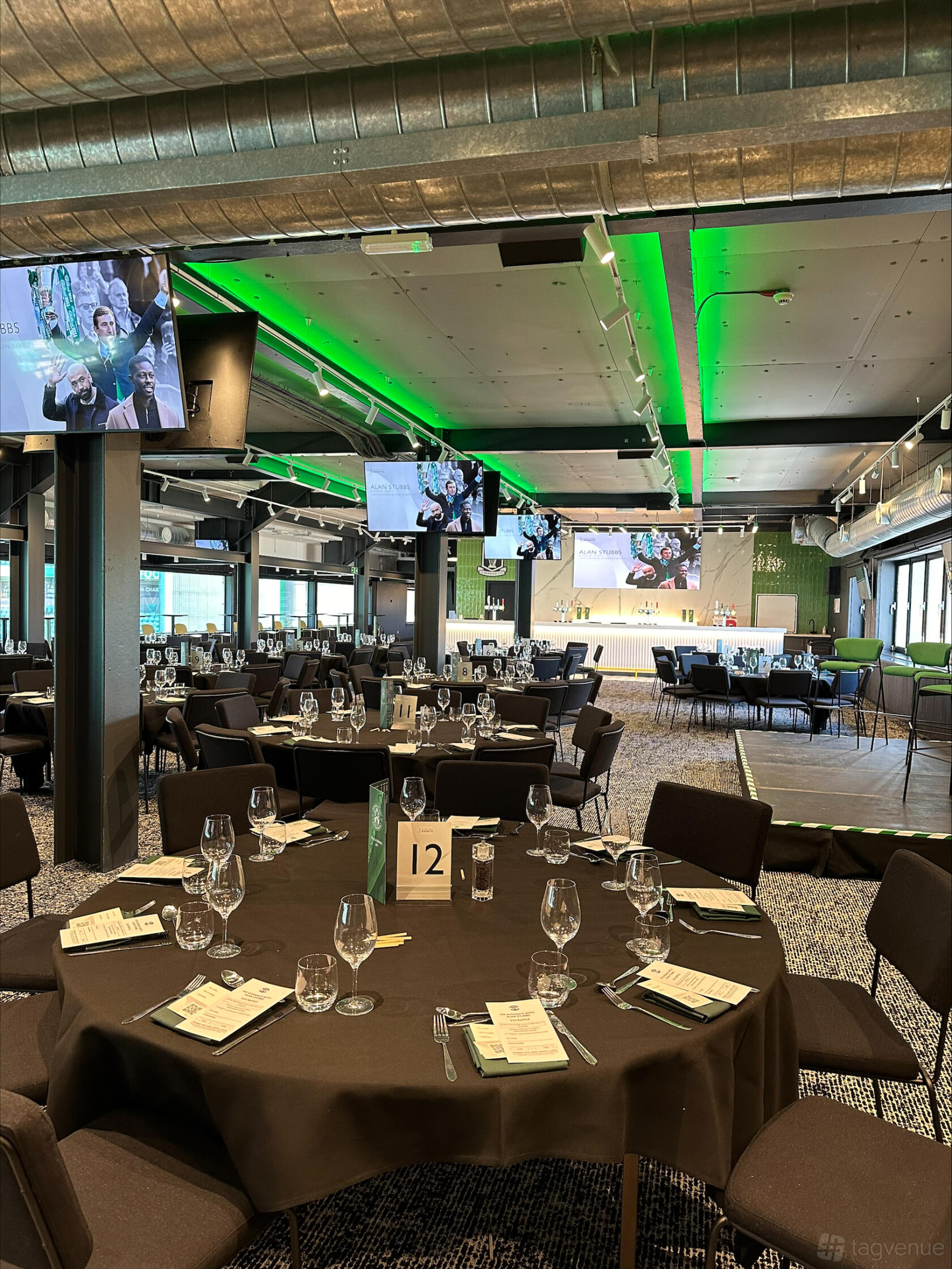 An event space with round tables set with glassware, stage, and TVs at Easter Road Stadium