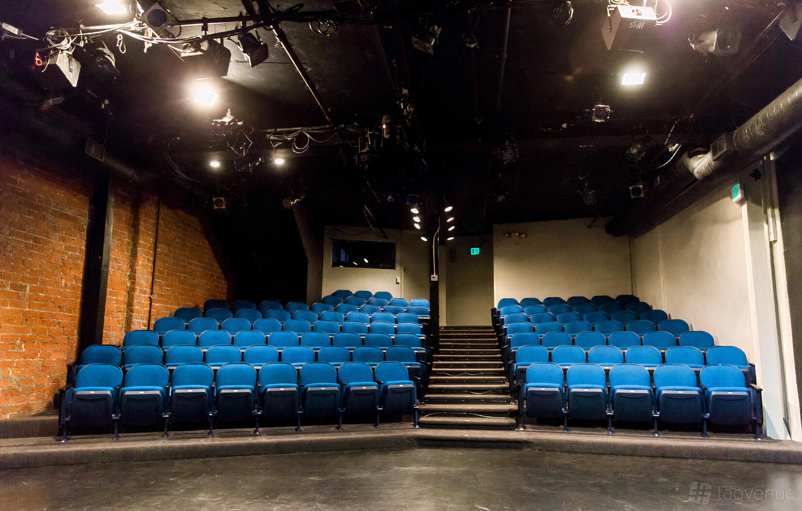 An auditorium with tiered rows of blue seats, exposed brick wall, and stage lighting at Hudson Theatres.