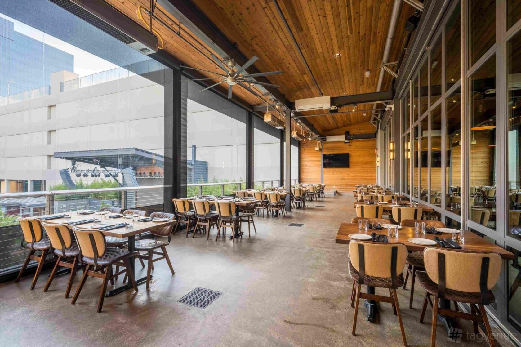 A covered terrace with wooden ceiling, ceiling fans, and rows of set tables overlooking an outdoor stage at Haywire Plano.
