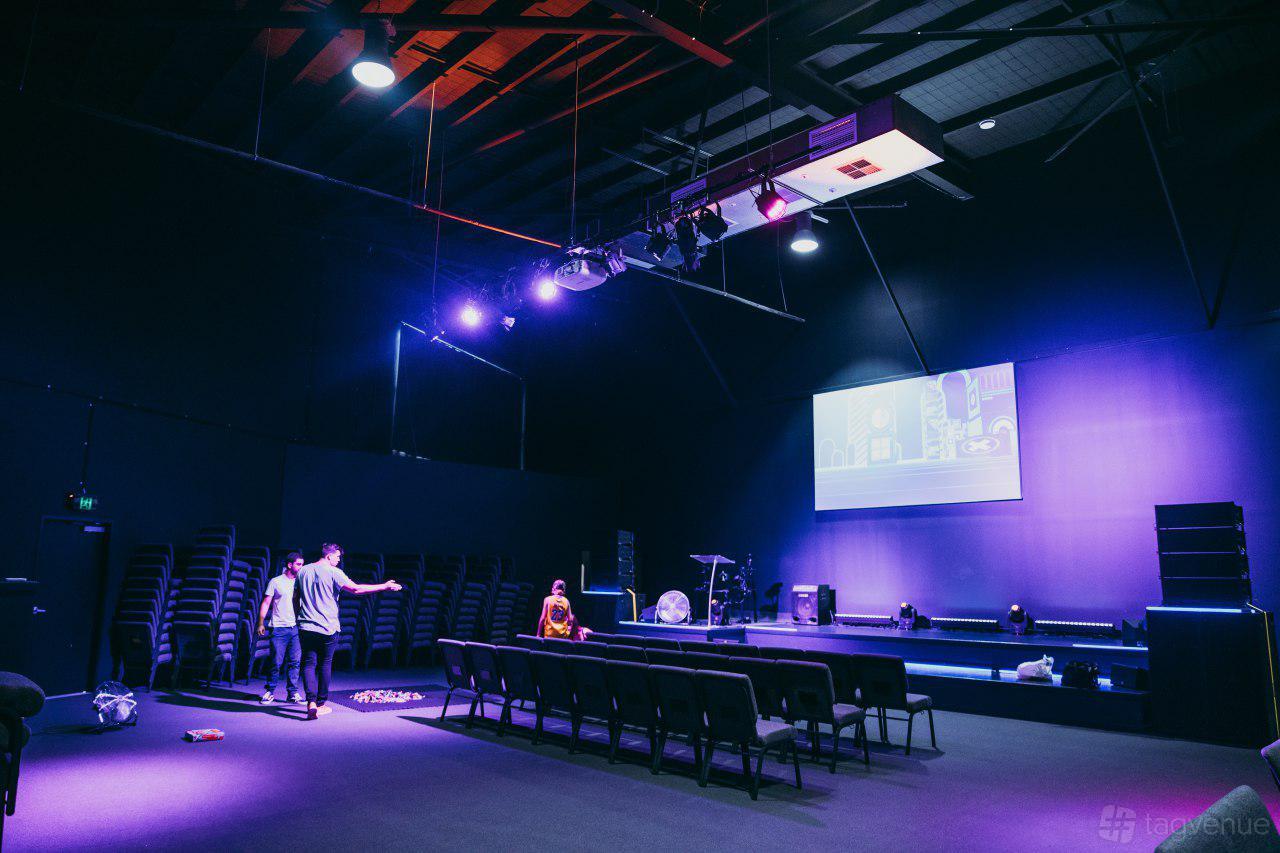 A conference centre with a raised stage, projector screen, and rows of black chairs at Glow.