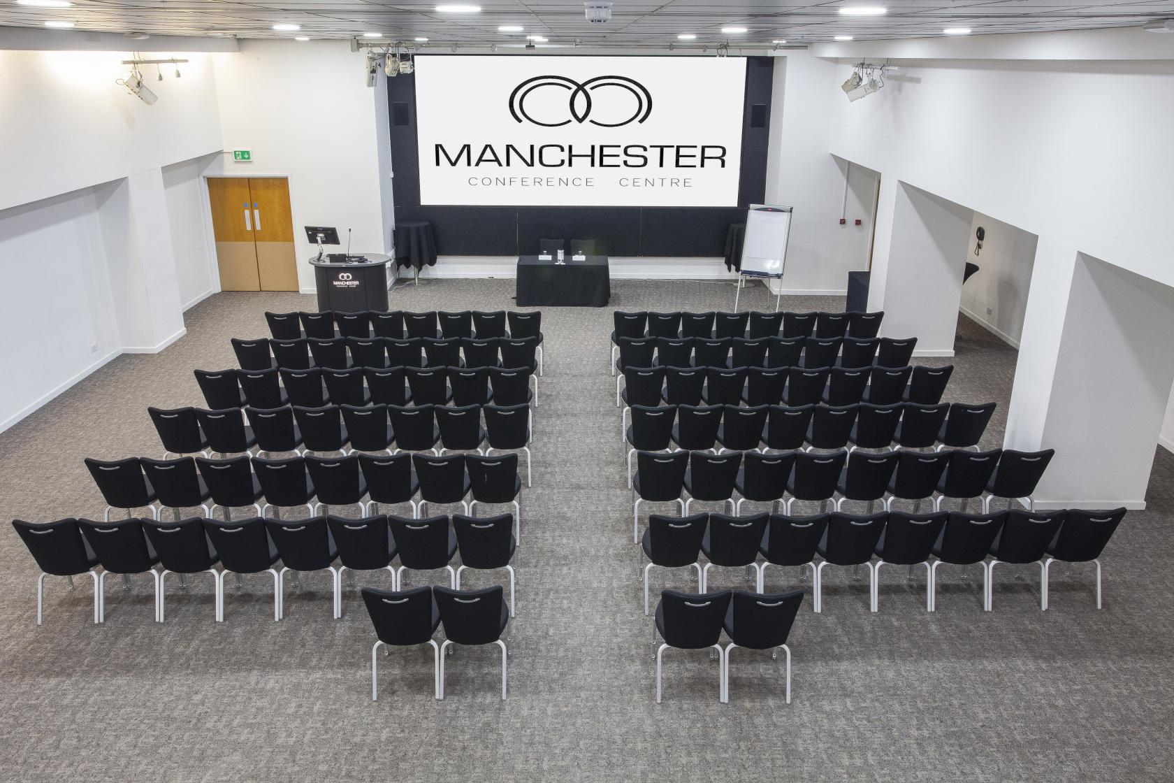An auditorium with rows of black chairs facing a stage with dual screens and podium at Manchester Conference Centre & Pendulum Hotel.