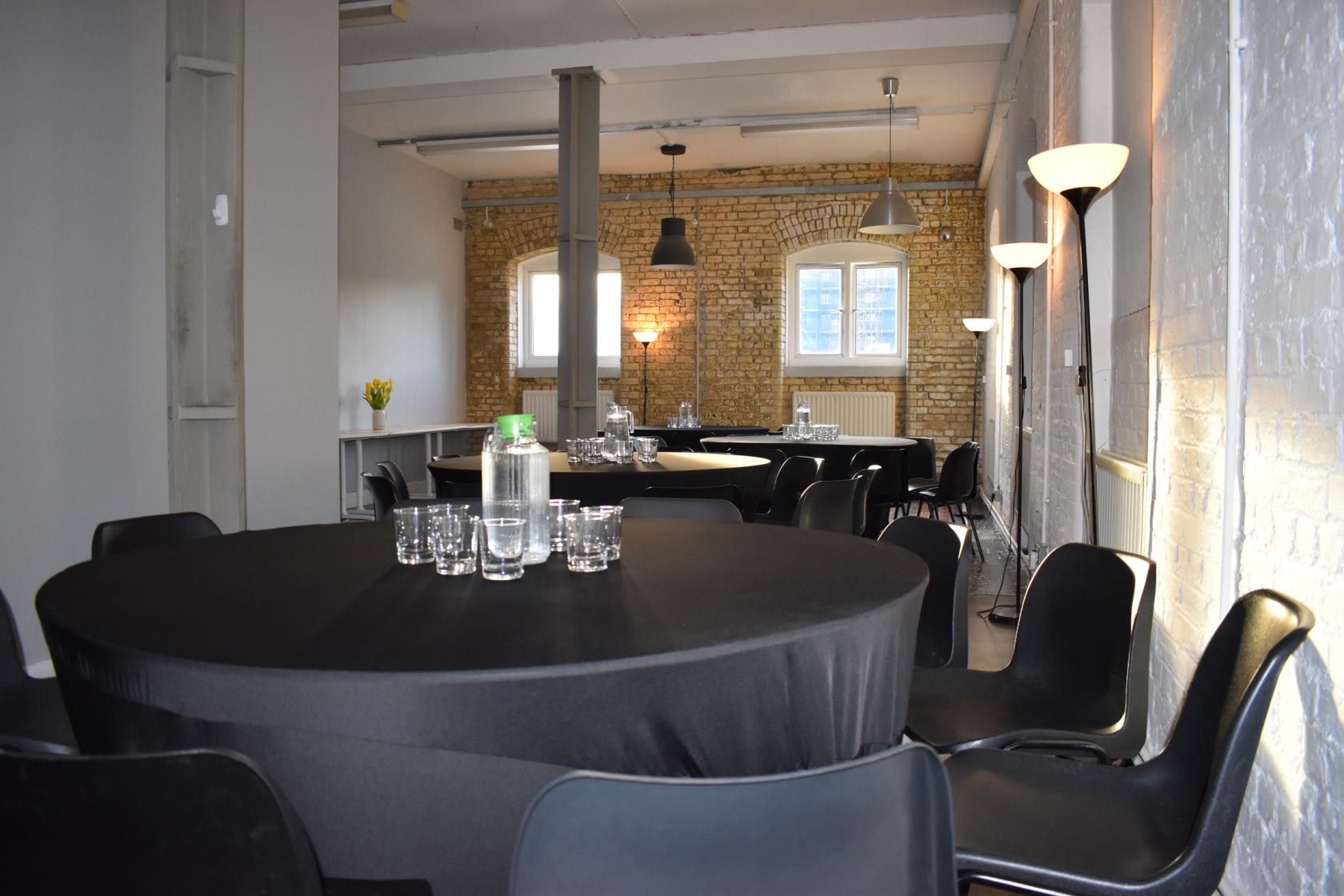 A function room with exposed brick walls, round tables covered in black linens, and industrial pendant lighting at Boathouse Studios, Barking.