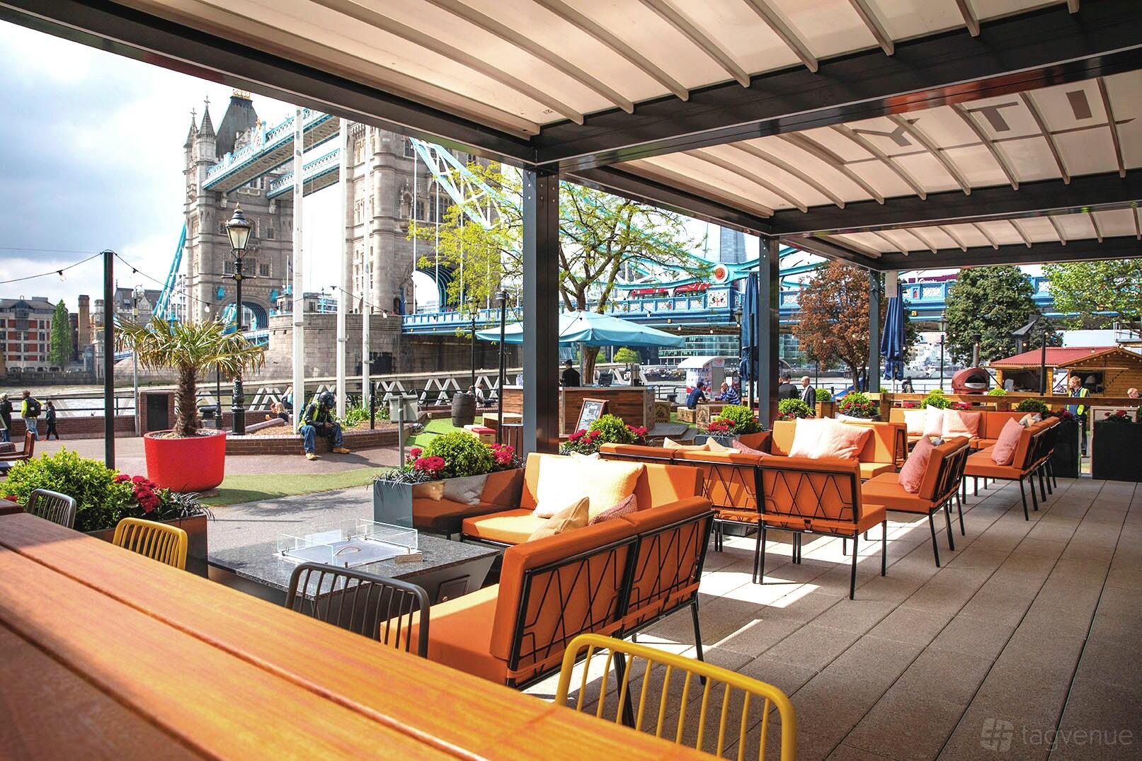 A riverside restaurant terrace with orange cushioned sofas, potted plants, and views of Tower Bridge at Vicinity.
