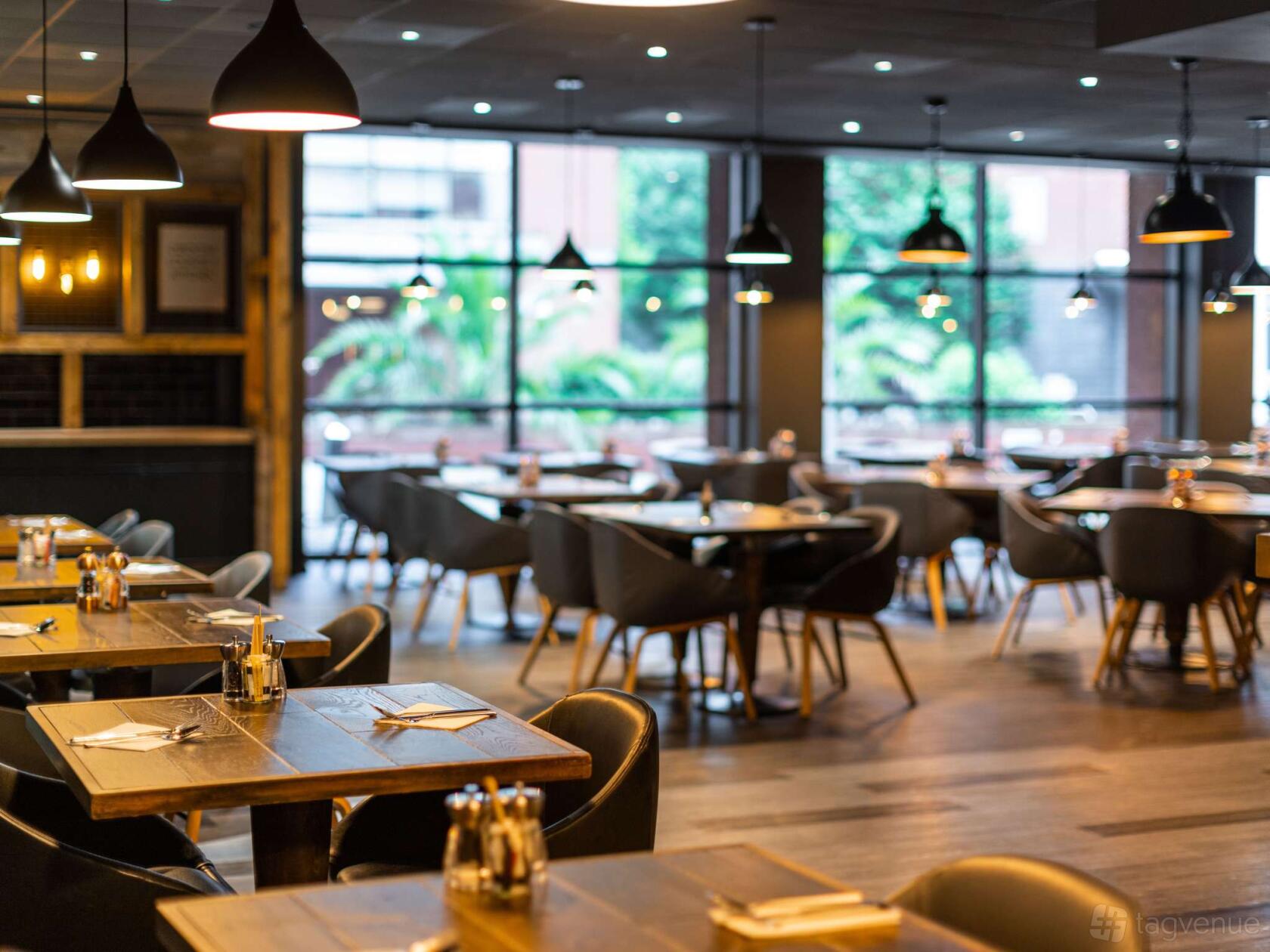 A hotel restaurant with floor-to-ceiling windows, wooden tables, and black chairs at Novotel Leeds Centre.