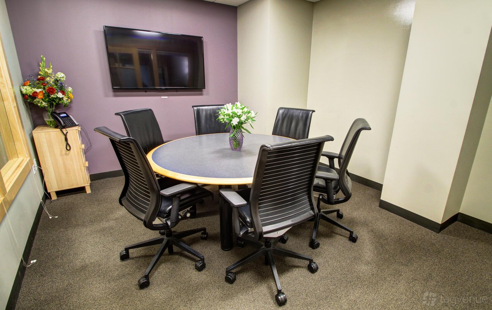 A meeting room with a round table, black office chairs, wall-mounted TV, and a bouquet of flowers at Intelligent Office.
