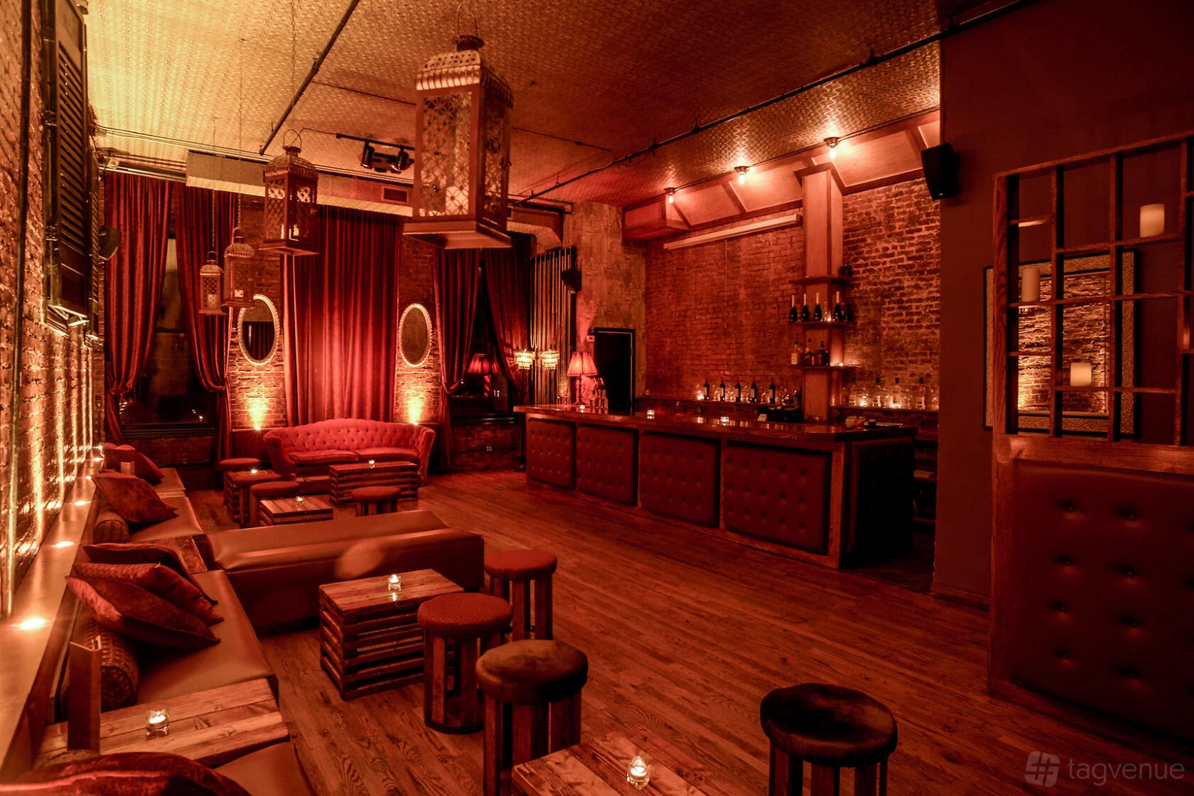 A bar with exposed brick walls, red velvet drapes, tufted seating, and pendant lanterns at Velvet Brooklyn.