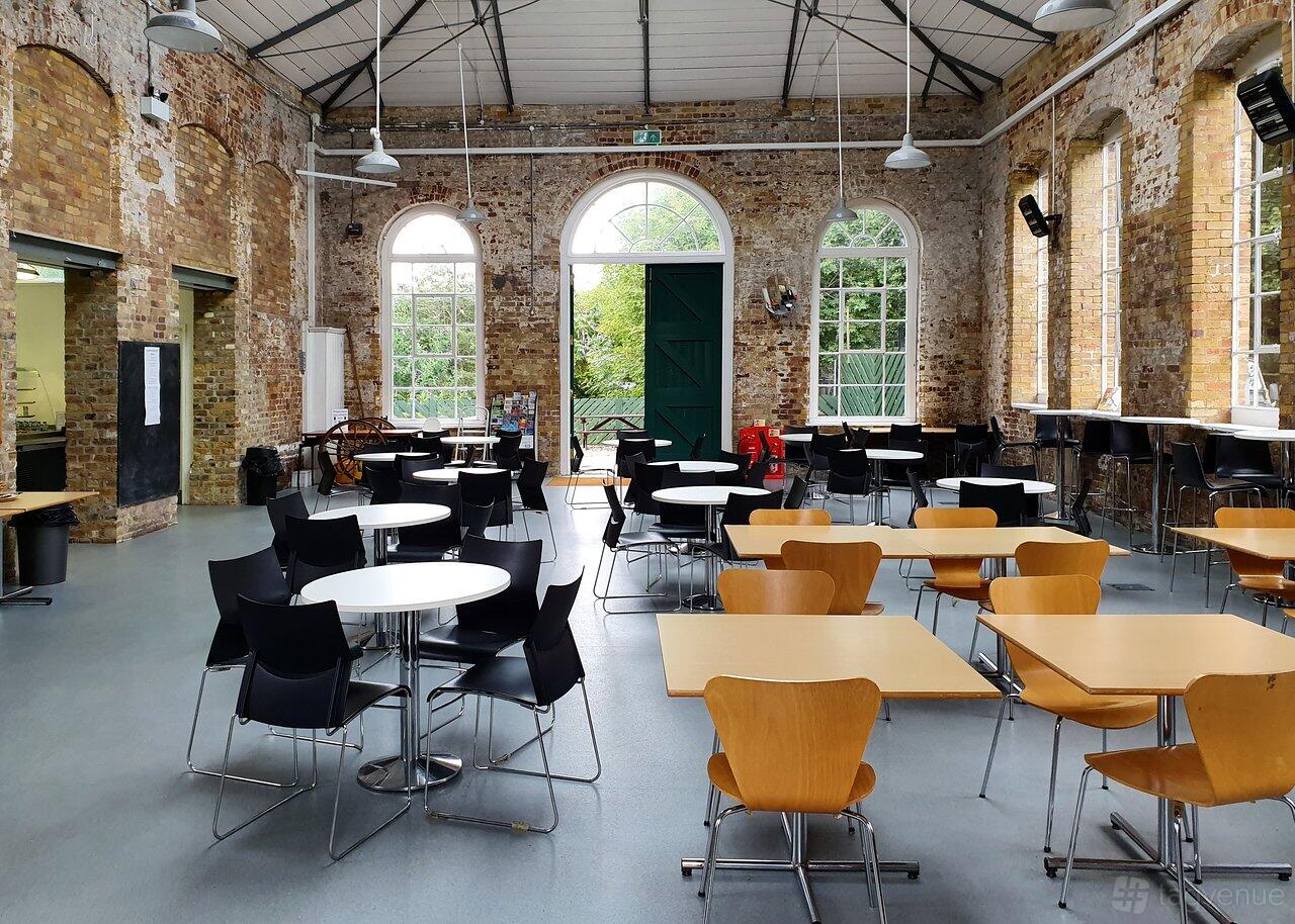 A cafe with exposed brick walls, high arched windows, and a mix of round and square tables at Royal Gunpowder Mills.