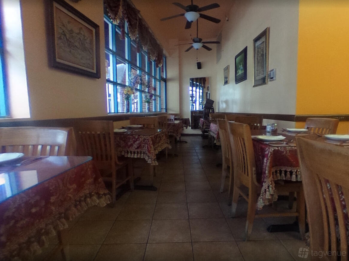 A restaurant dining room with wooden chairs, patterned tablecloths, framed art, and large windows at Shamshiri Restaurant.
