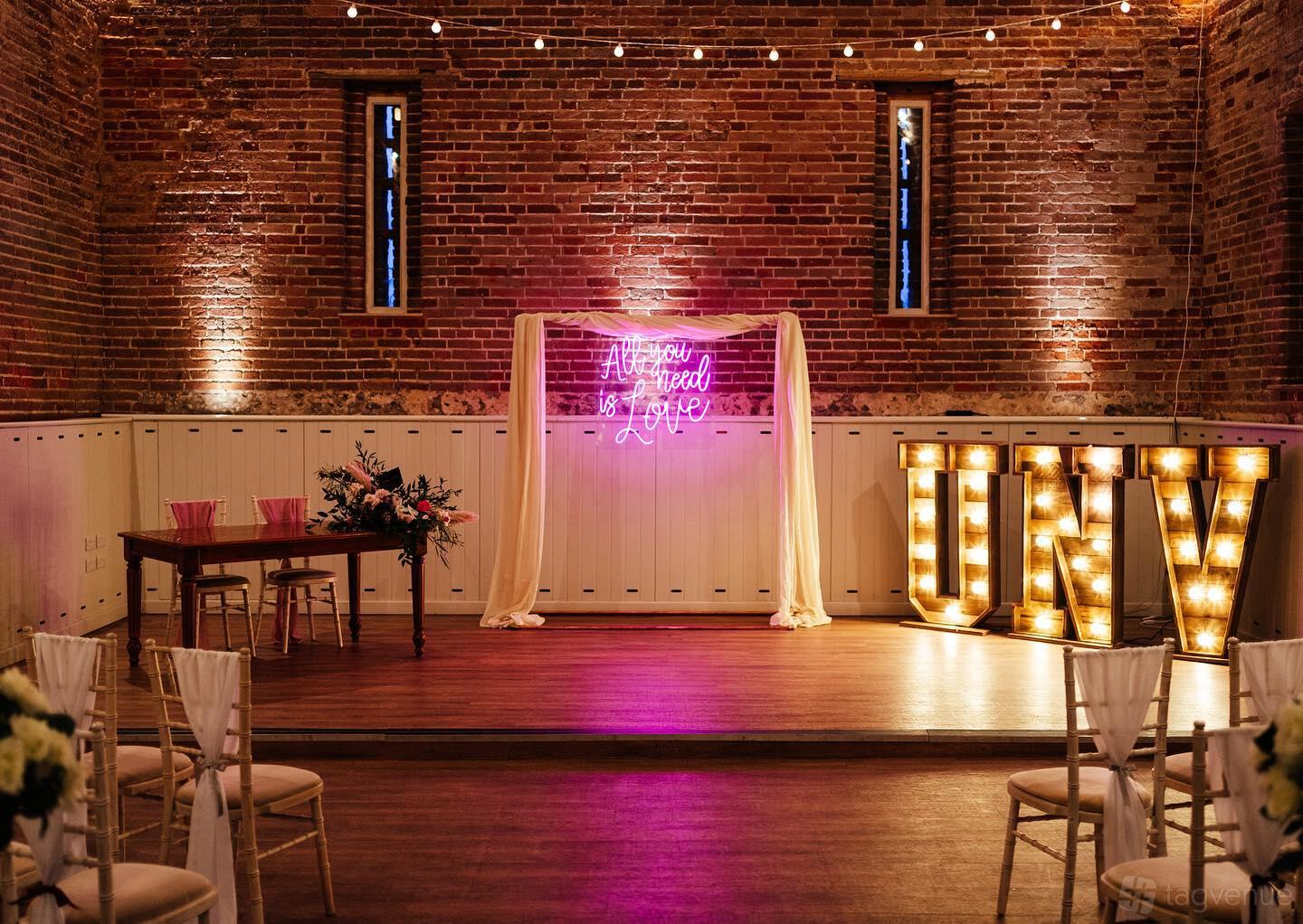 A barn with exposed brick walls, string lights, and a raised stage with neon and marquee signage at Manor Mews.