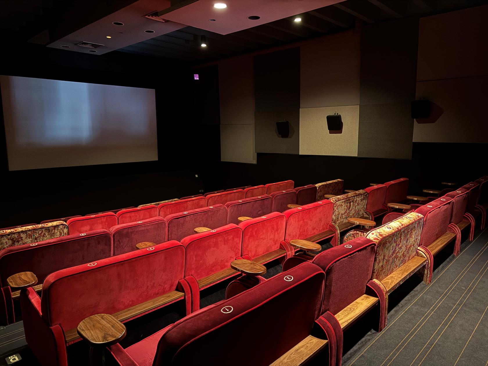 An event space with red velvet cinema seats, wood armrests, and a large screen at Everyman Walton.