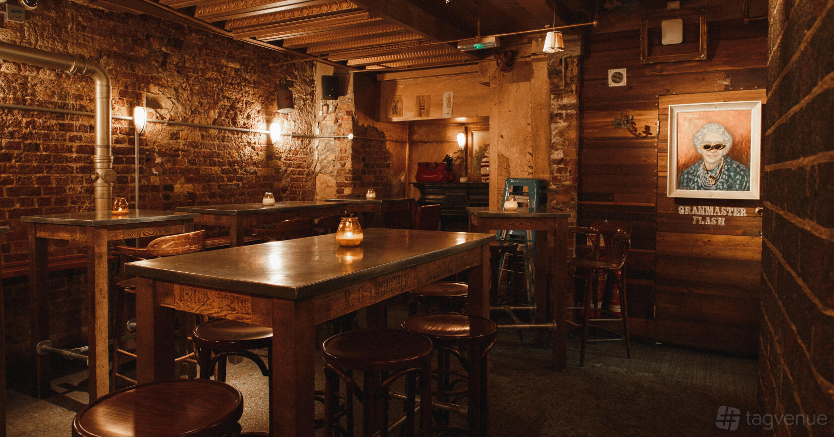 A cocktail bar with exposed brick walls, wooden tables, warm lighting, and a quirky portrait at The Breakfast Club Spitalfields.
