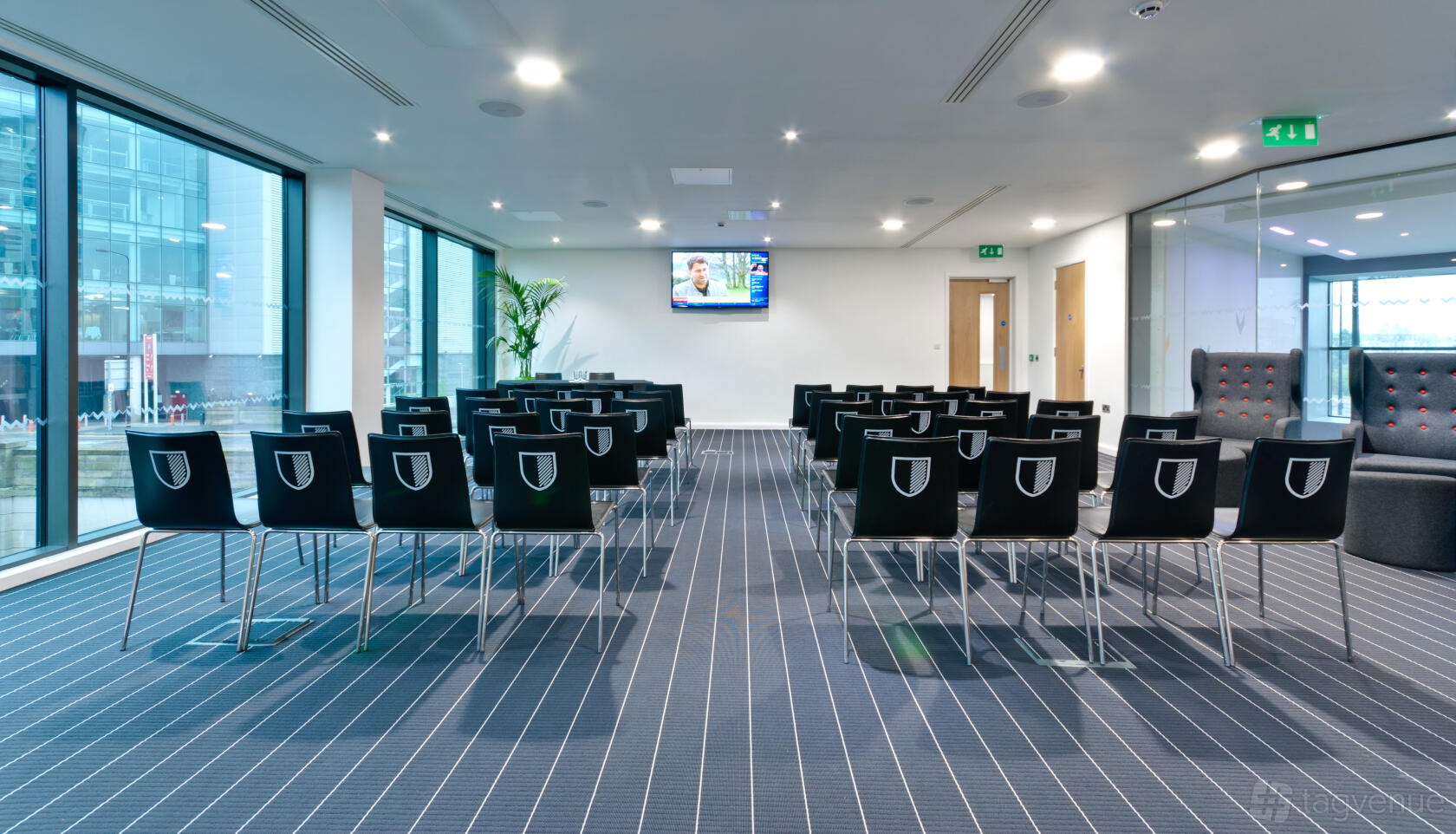 A lounge with rows of black chairs facing a wall-mounted TV, floor-to-ceiling windows, and striped carpeting at Hotel Football.