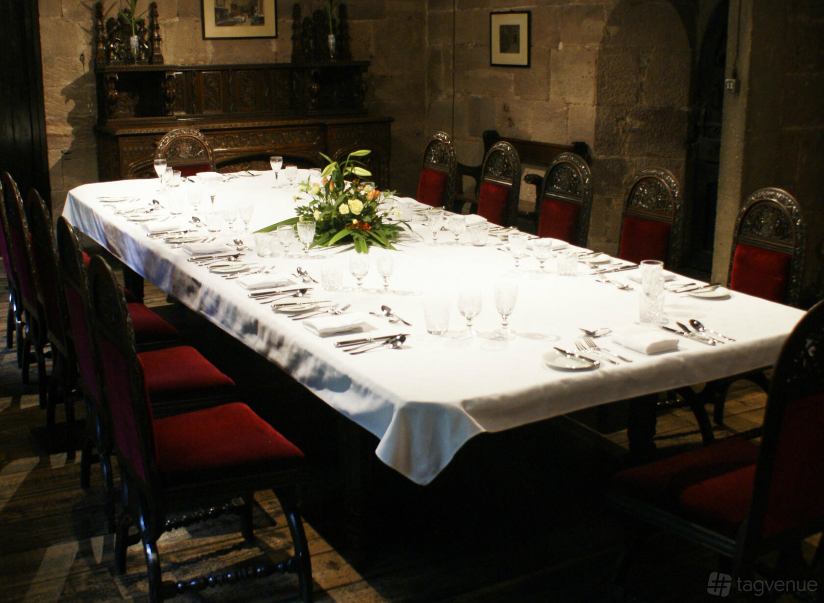 A historic dining room with a long table set with white linens, red upholstered chairs, and floral centerpiece at Chetham's Library.