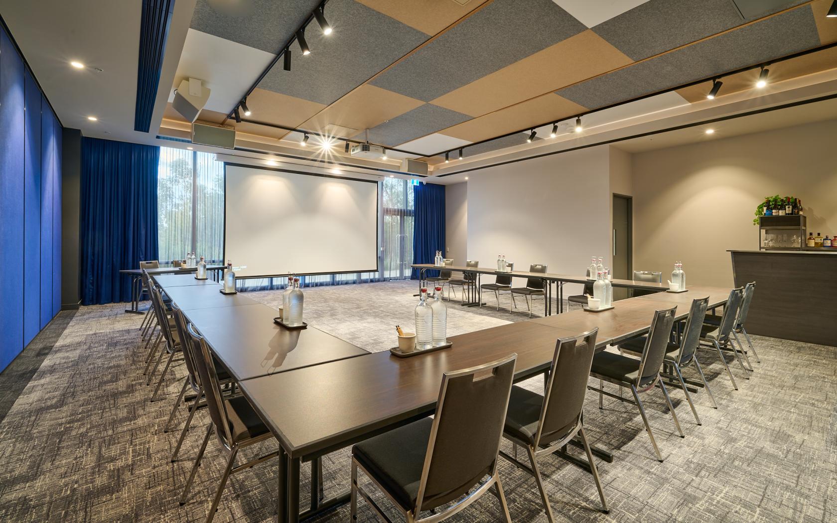 A hotel meeting room with U-shaped tables, grey chairs, floor-to-ceiling windows, and a ceiling with acoustic panels at Mantra Melbourne Epping.