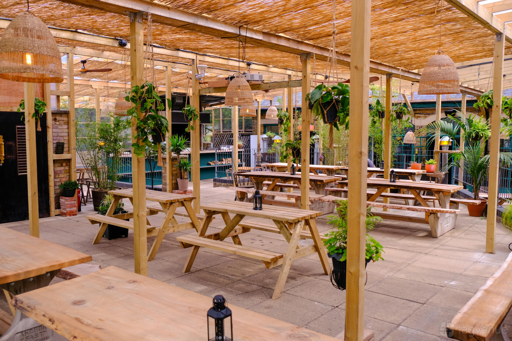 A pub terrace with wooden picnic tables, hanging woven lamps, and potted plants at Pacific Tavern.