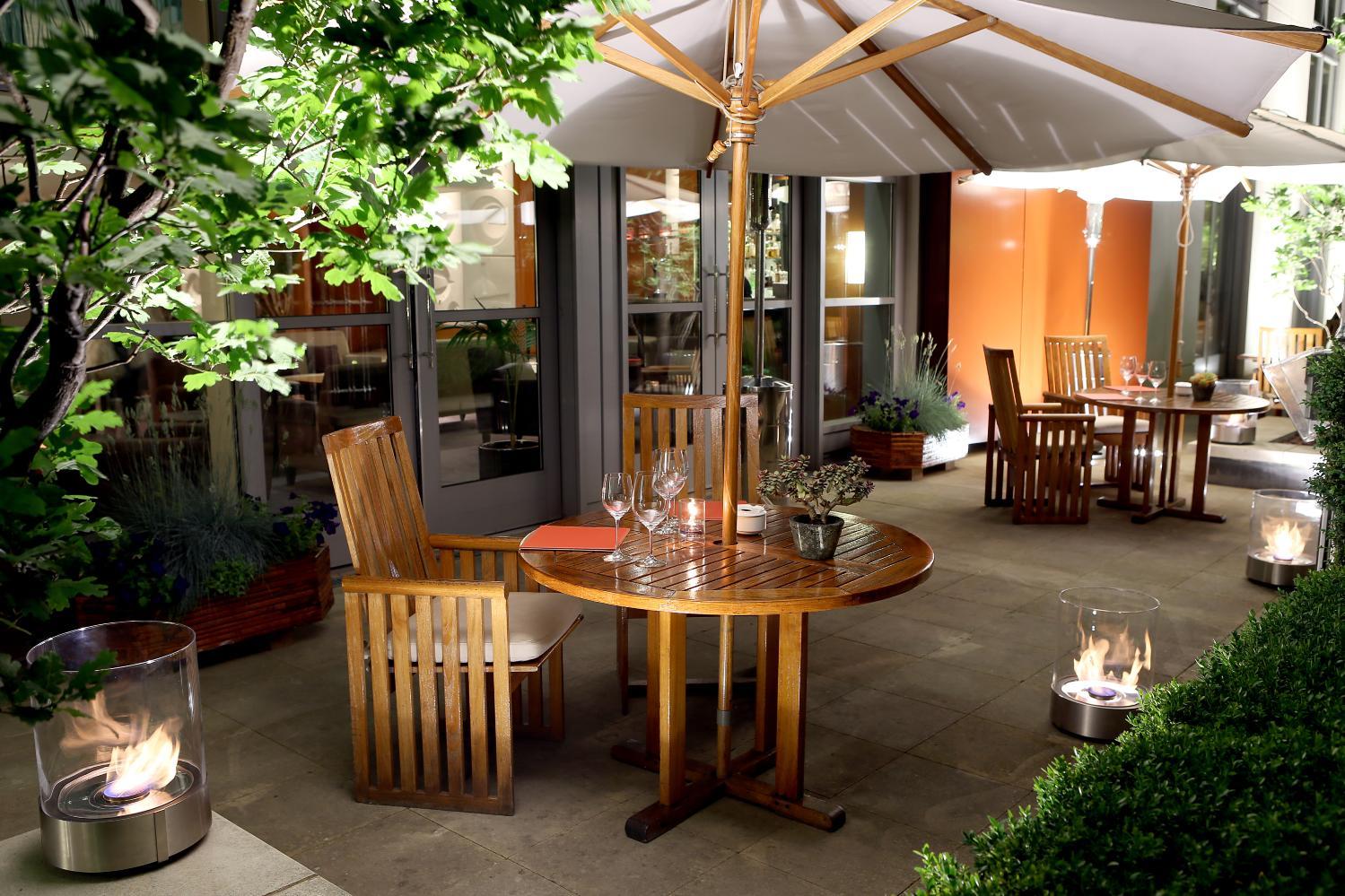 An outdoor hotel terrace with wooden tables and chairs, large umbrellas, and glass-enclosed fire pits at Canary Riverside Plaza Hotel.