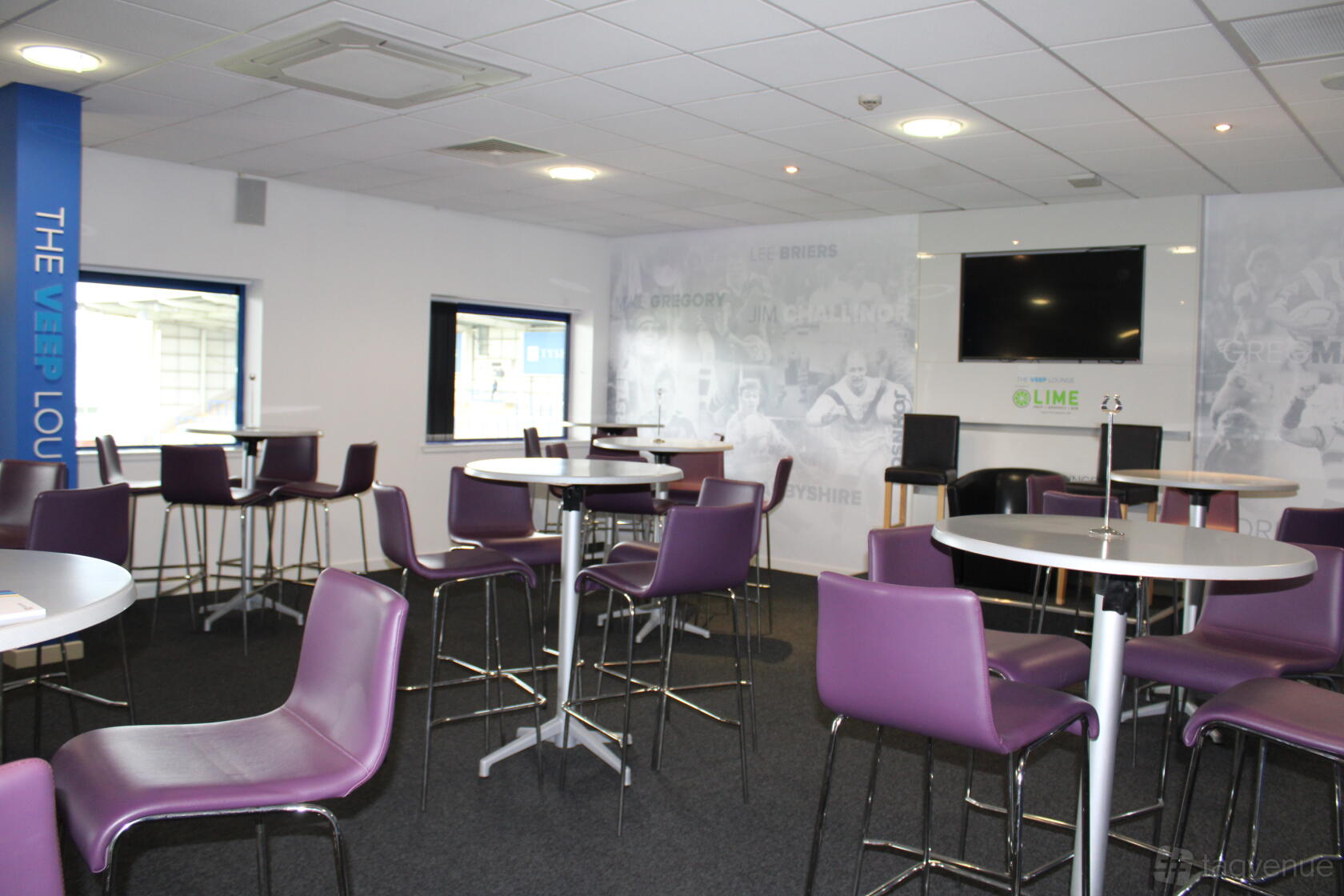 A stadium lounge with round high-top tables, purple bar stools, large windows, and a wall-mounted TV at The Halliwell Jones Stadium.