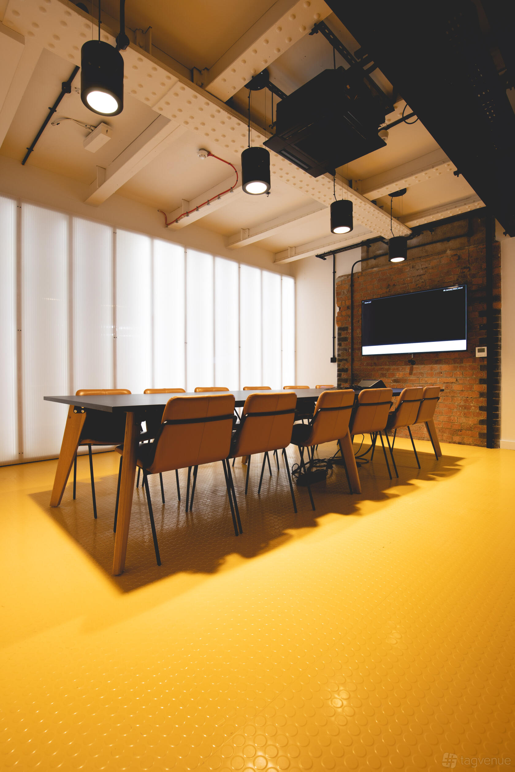 A meeting room with a long table, tan chairs, exposed brick wall, and wall-mounted screen at Department Bonded Warehouse.
