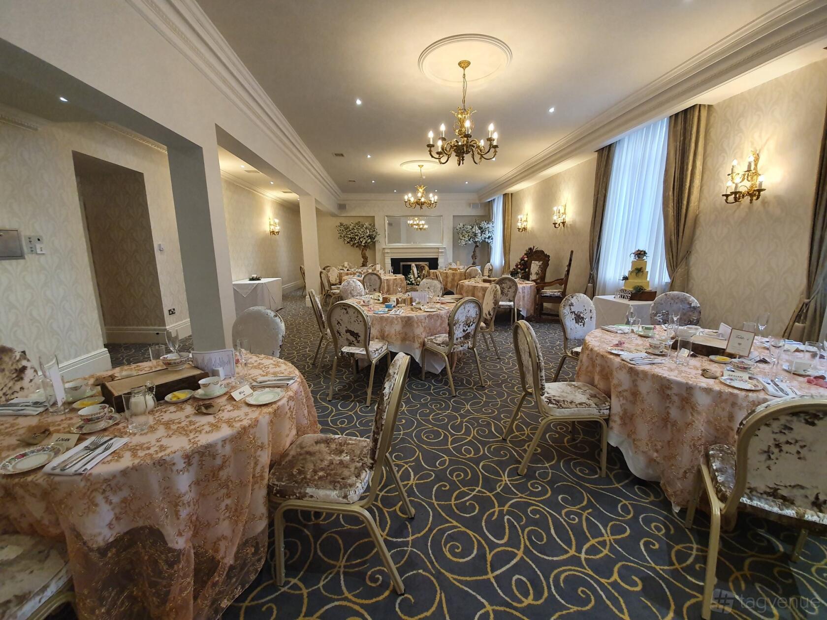 An event space with round tables draped in floral linens, plush chairs, chandeliers, and a fireplace at Grand Hotel Tynemouth.