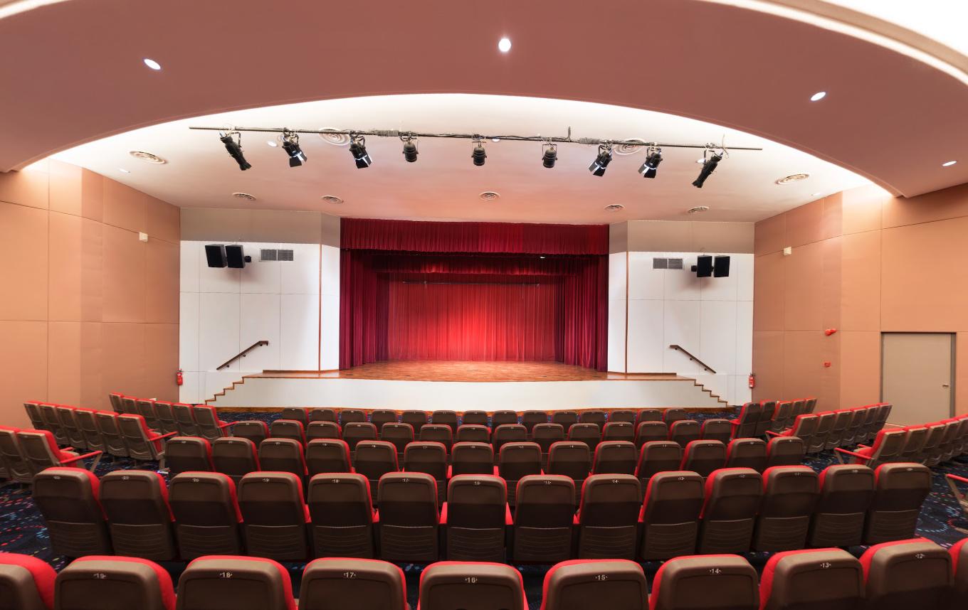 An auditorium with tiered seating facing a large stage, red curtains, and overhead stage lighting at The Chevrons.