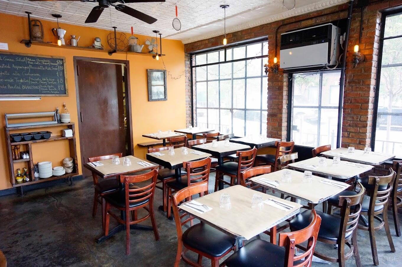 A restaurant with large windows, exposed brick walls, chalkboard menu, and rows of wooden tables at Testo.