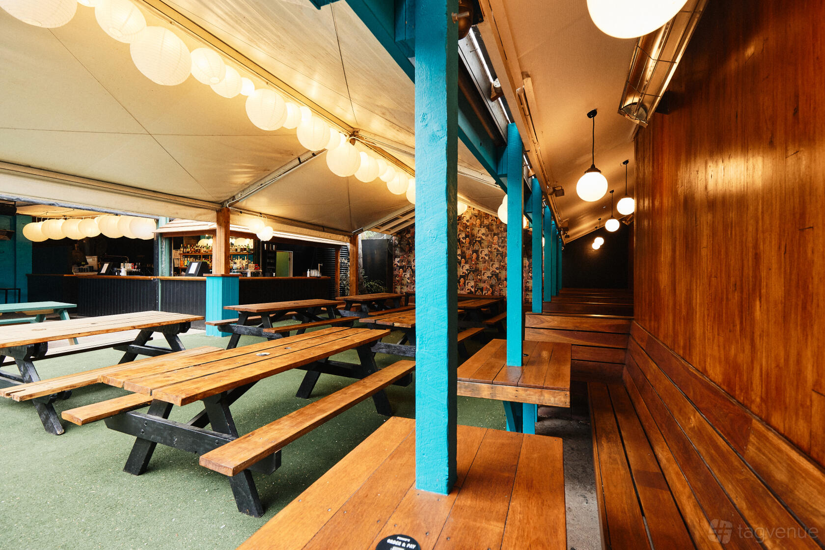 A covered beer garden with long wooden benches, globe pendant lights, and an artificial grass floor at The Penny Black.