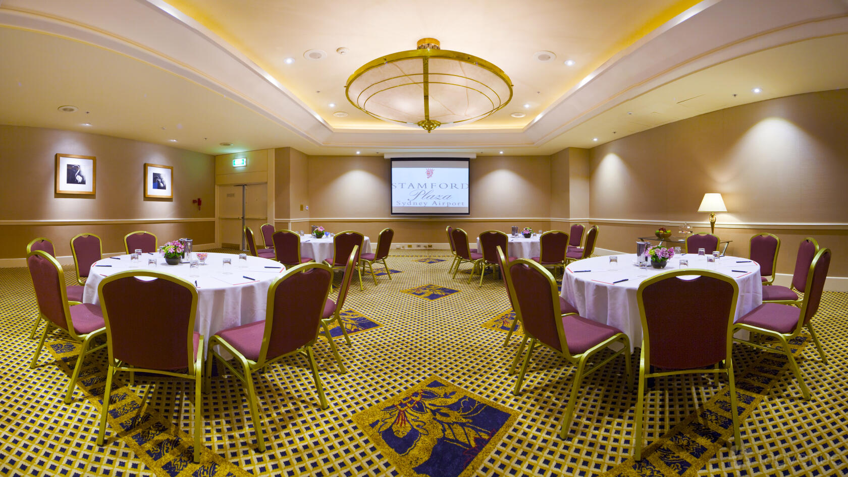 A meeting room with round tables, red chairs, a ceiling chandelier, and a projector screen at Stamford Plaza Sydney Airport Hotel.