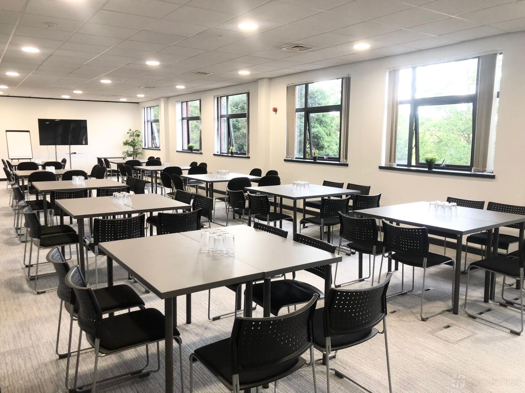 A meeting room with rectangular tables, black chairs, large windows, and ceiling lights at Lawrence House - Hub26.