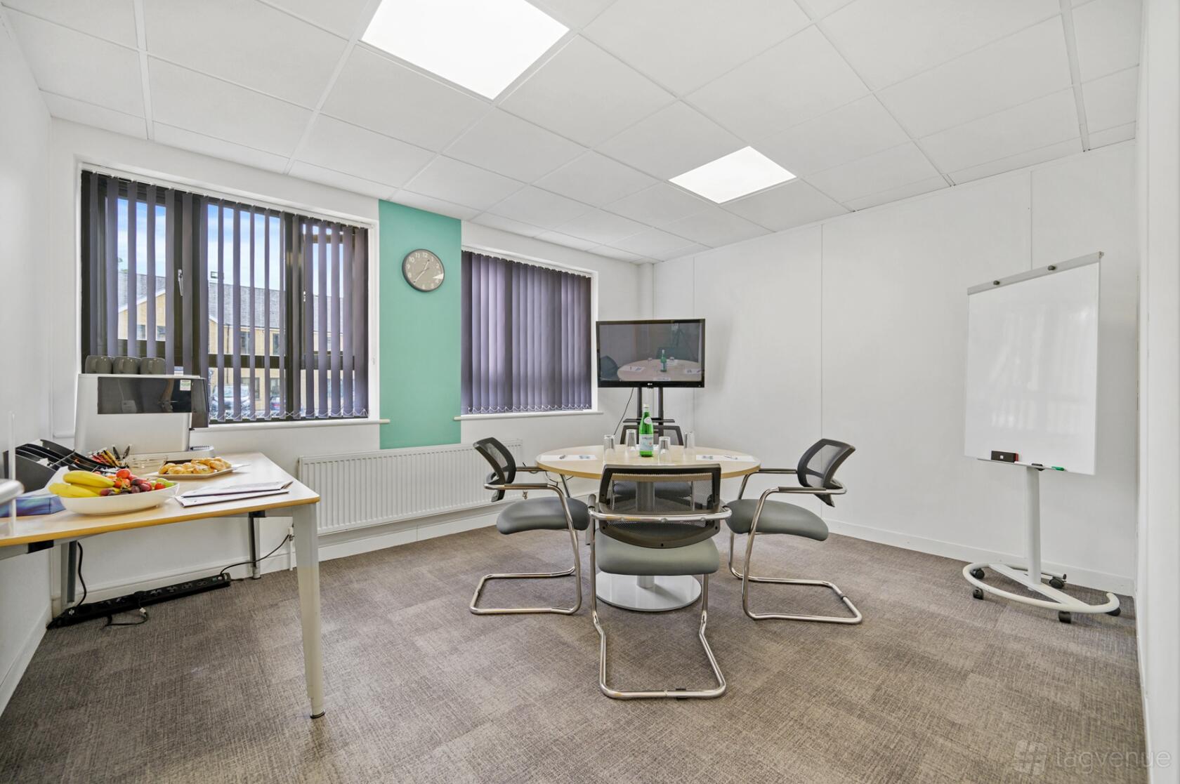 A meeting room with a round table, four chairs, wall-mounted screen, flip chart, and snack table at UBCUK Cirencester Office Park.