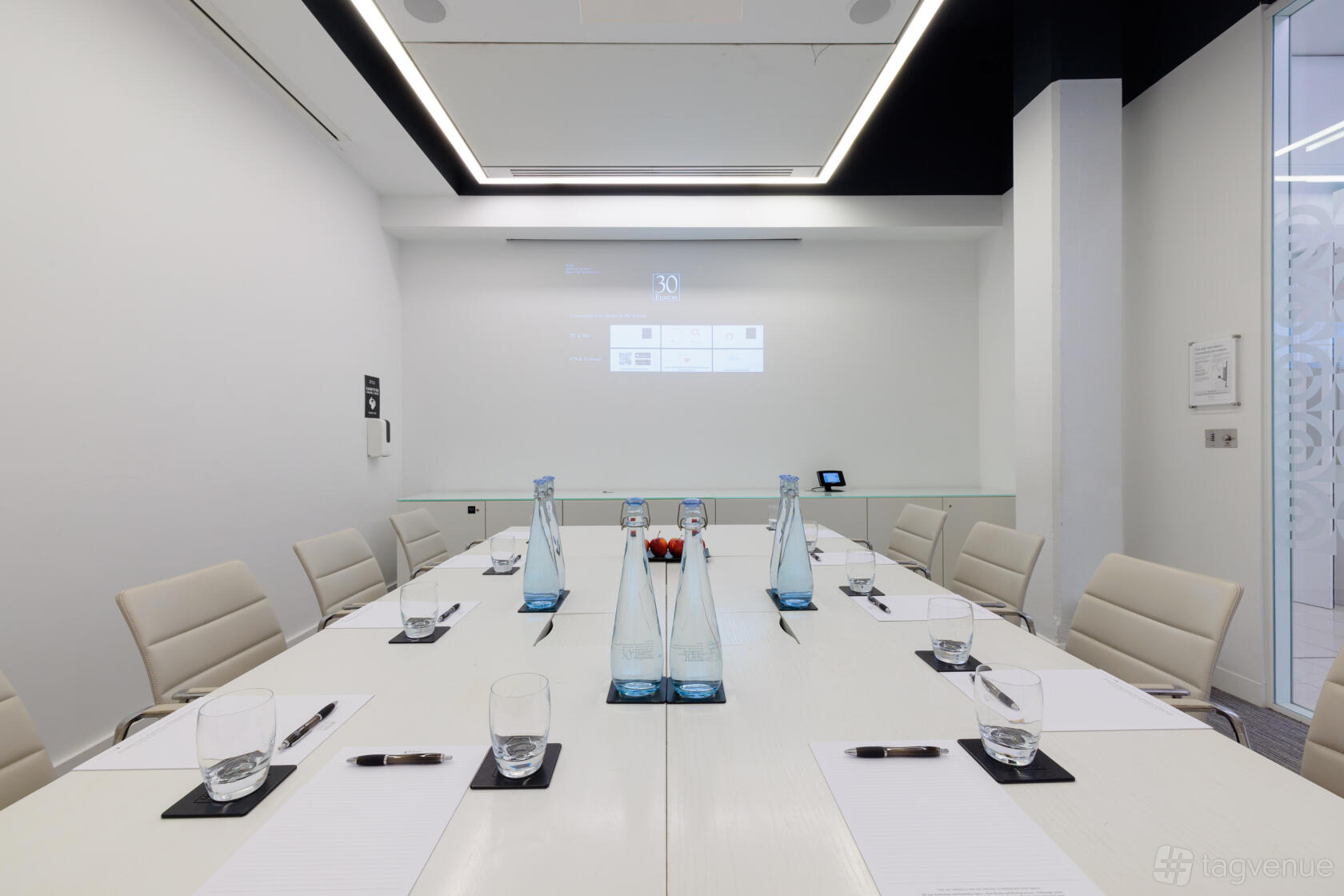 A meeting room with a long conference table, white leather chairs, glass water bottles, and a wall-mounted screen at 30 Euston Square.